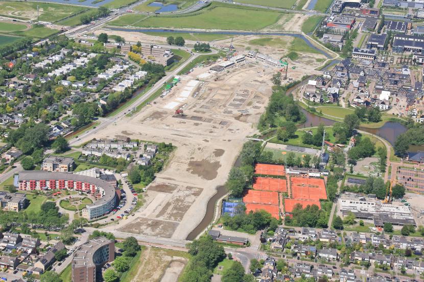 Luchtfoto van een bouwterrein waar een aannemer woningen bouwt. Links en rechts woningen en een tennisbaan. Op de achtergrond een singel.